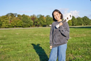 Güzel Koreli kız ukulele ile poz veriyor, genç müzisyen güneşli bir günde yeşil parkta oynuyor..