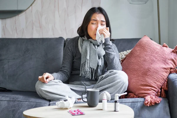 Portrait of korean woman sneezing, feeling sick, staying at home with flu or cold, neck wrapped with scarf, staying at home, taking medication.