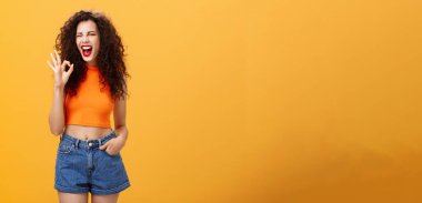 Harika bir parti. Neşeli, coşkulu, kıvırcık saçlı, kırmızı rujlu, stil sahibi, üstü açık, heyecanlı, gülümseyen ve turuncu arka planda mükemmel bir işaret veren genç bir kadın..