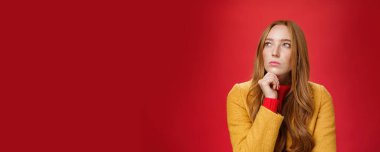 Kararlı ve odaklanmış yaratıcı düşünceli kızıl saçlı kadının sol üst köşeye bakıp çeneye dokunurken, düşünürken, seçim yaparken ya da kırmızı arkaplan üzerinden bilgileri hatırlarken yakın plan çekimi..