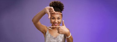 Yaratıcı, parlak elbiseler içinde, ilham arayan Afrikalı bir kadın parmak çerçevesini mutlu eden, gülümseyen, mükemmel bir fotoğraf açısı bulan, mavi arka planda duran,.