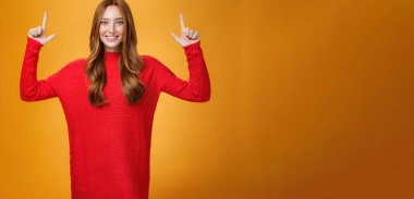 Hevesli ve heyecanlı kızıl saçlı, kırmızı, sıcak kazaklı kadın yükselen elleriyle işaret ediyor ve büyük bir heyecanla gülümsüyor, turuncu arka planda şaşkın hissediyordu..