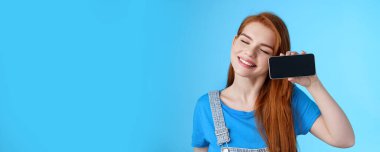 Tasasız, hassas, Avrupalı bir kadın, gülümseyen, kapalı gözlü, akıllı telefonu yatay olarak tutan, sevgili fotoğrafları gösteren, romantik videolar gösteren, neşeli mavi arka planda duran..