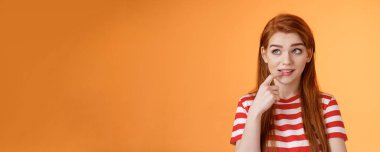 Tuhaf, sevimli, kızıl saçlı kız arkadaş, aptal ısıran parmak, ilgili sol köşesine bak, merak uyandırıcı bir fikir, kafa yorma, karar verme sırası online, düşünceli turuncu arka plan.
