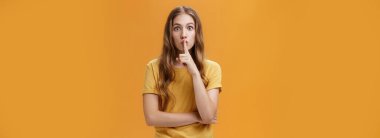 Oyun sırasında sessiz ol. Ciddi ve kararlı, dalgalı saçlı, sessiz jest yapan, ağzı işaret parmaklı, sessiz kalan, turuncu duvarda sır saklayan genç bir kadın..