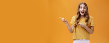 Heyecanlı, şaşırmış ve hayrete düşmüş genç Avrupalı zayıf kadının portresi. Dalgalı doğal saç stiliyle çenesini düşürüyor. Solu göstererek turuncu duvarın ardında harika bir şey hakkında sorular soruyor..