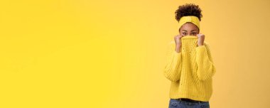 Yakışıklı, aptal, Afro-Amerikan modern kızı. Kazağını yüzüne geçirip kamerayı gizliyor. Gülümsüyor, saklambaç oynuyor ve sarı arka planı taklit ediyor..