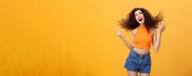 Aptal pozitif ve enerjik bir kadın portresi. Kıvırcık saçlı, kıvırcık saçlı, saçlarını savurup gülümseyen, sağ üst köşede büyüleyici bakışları olan turuncu duvarın üzerinde mutlu poz veren..