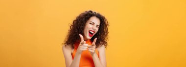 N 'aber? Arkadaş canlısı ve cana yakın kıvırcık saçlı, kırmızı rujlu, parmak tabancalarıyla kameraya doğrultulmuş bir kadın. Sanki turuncu arka planda arkadaşına göz kırpıyormuş gibi..