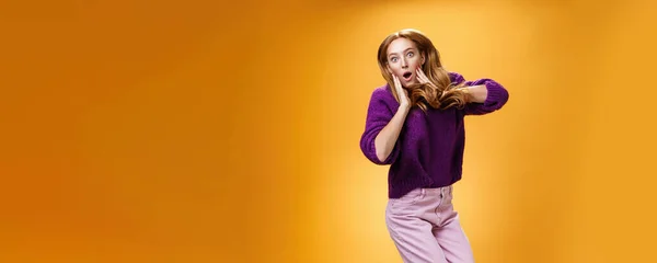 Lifestyle. Impressed and surprised amazed good-looking redhead female in purple sweater open mouth astonished and holding hands on cheeks as jumping popping eyes amused over orange background.