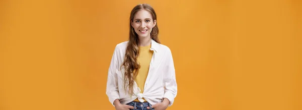 Lifestyle. Stylish young female designer wants help friend shopping holding hand in pockets smiling joyfully and self-assured at camera wearing trendy blouse over yellow t-shirt posing over orange