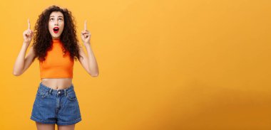 Şaşırmış duygusal ve karizmatik genç, kıvırcık saçlı, kırmızı rujlu bir kadının portresi turuncu arka planda etkilenmiş ve hayretler içinde kalmış. Boşluğu kopyala