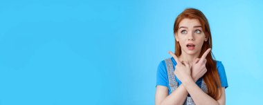 Belirsiz kızıl saçlı kadın karar veriyor, çapraz kollu, sağ üst köşeleri işaret ediyor, tereddütlü, düşünceli, açık ağızlı, ürünleri seçiyor, mavi arka plan.