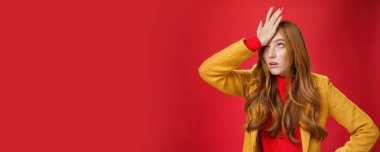 Redhead businesswoman making facepalm gesture with hand on forehead rolling eyes up from annoyance and irritation as being shocked with how dumb client is sighing bothered over red wall, tired.