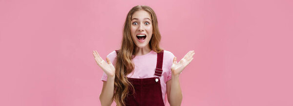 Waist-up shot of excited happy and impressed charming caucasian girlfriend in overalls and t-shirt raising hands in surprise and amazement cheering smiling broadly from astonishment over pink wall
