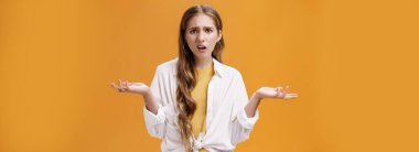 What, I do not understand. Portrait of arrogant questioned impolite girl being pissed with questiones shrugging with hands spread sideways making irritated grimace standing confused over orange wall.