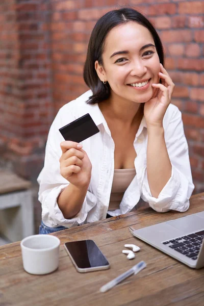 Beautiful young asian woman with credit card, sitting near laptop and smiling, paying bills, shops online, order smth on computer.