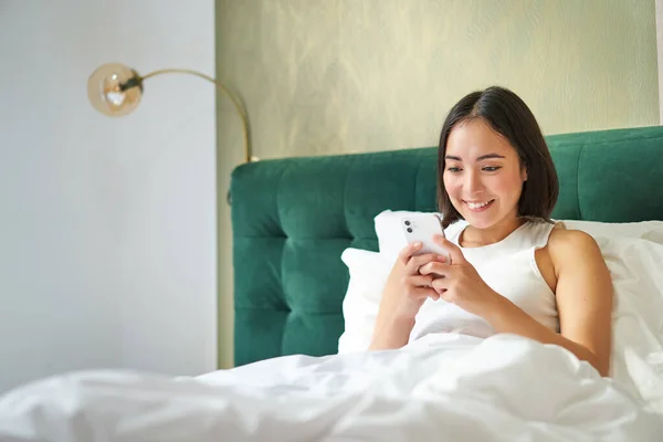 Beautiful smiling asian woman in bed, reading message on smartphone and laughing, enjoying lazy day in bedroom, using mobile phone.