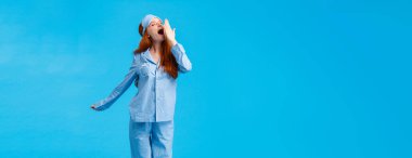 Ne harika bir sabah. Tam boy dikey stüdyo, esneme, uyku maskesi ve pijama giymiş, esneme ve kapağı açık, mavi arka planda, gözleri kapalı, tatlı, beyaz saçlı bir kız..