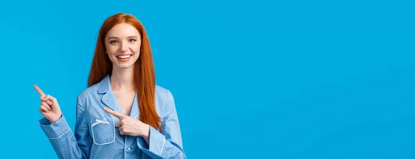 Friendly attractive redhead female student showing person way in dormitory college, pointing right and smiling wearing nightwear, going sleep, showing her daily skincare routine, blue background.