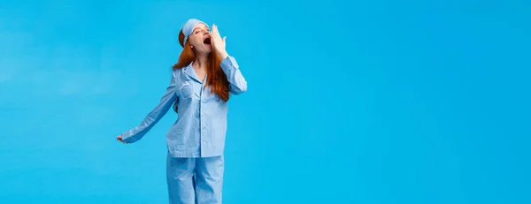 What wonderful morning. Full-length vertical studio shot yawning cute caucasian redhead girl in sleep mask and pyjama, stretching and cover opened mouth with closed eyes, blue background.