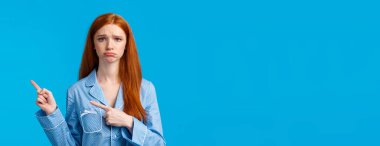 Pişmanlık, kötü ruh hali ve reklam konsepti. Üzgün, mutsuz, gece elbisesi giymiş, somurtan, somurtan, hayal kırıklığı ya da kıskançlık gösteren, parmaklarını sağa doğru gösteren, mavi arka planda duran mutsuz bir kızıl kız..