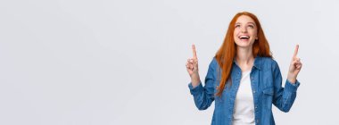 Kot gömlekli neşeli, karizmatik kızıl kadın, mutlu bir gülümsemeyle bakıyor ve işaret ediyor, gülüyor, tam olarak neye ihtiyacı olduğunu buluyor, rüya görüyor, beyaz arka plan pozitif duygularla ayakta duruyor..