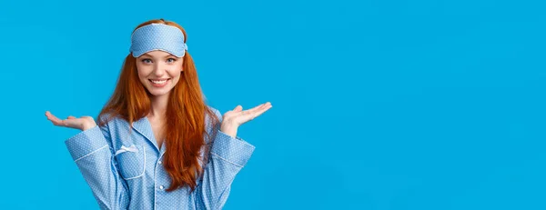 Who knows, dont care. Carefree and unbothered happy smiling redhead female in nightwear, sleep mask, shrugging indecisive and clueless with hands raised sideways, standing blue background.