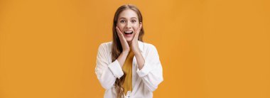 Heyecanlı ve keyifli, zarif, modaya uygun kıyafetleri olan, şaşırmış ve heyecanlı bakışları olan, turuncu duvarın üzerinde poz veren, harika haberleri dinlemekten zevk alan genç bir kadın. Vücut