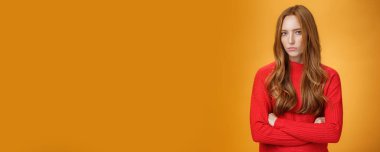 Pasif agresif poz veren şüpheli, gergin ve savunmacı kızıl kız. İnançsız bir şekilde kameraya bakıyor ve küçümsüyor. Turuncu arka plana alınıyor. Vücut dili