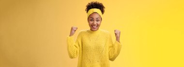 Şaşırmış sevimli Afro-Amerikalı kız piyango ikramiyesini kazandığına inanamıyor. Yumruklarını sallıyor, gülümsüyor, geniş gözlere gülüyor başarıyı kutlarken hayran kalıyor, zafer sarısı arka planda..