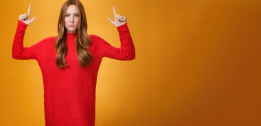 Aşırı hayal kırıklığına uğramış, sıkıntılı, 20 'li yaşlarda, kızıl saçlı, kırmızı, kalın kazaklı bir kadın. Üzüntüden çatık kaşlı, turuncu arka planda memnuniyetsizliğe işaret eden ellerini kaldırıyor..