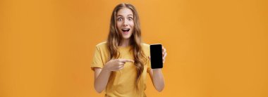 Harika akıllı telefon özellikleriyle şaşırmış bir kız. Uygulamada cep telefonunun cihaz ekranını işaret ettiğini gösteren dalgalı doğal saç stiline sahip neşeli ve hayret verici Avrupalı ince kadının portresi. Boşluğu kopyala