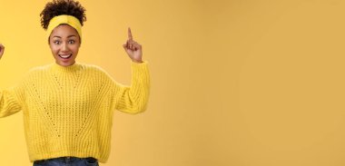 Süveter tokalı, etkileyici bir para kazanan, etkileyici duygusal, Afro-Amerikalı genç bir kadın işaret parmaklarını göstererek şaşırtıcı bir şekilde sarı arka planda sırıtıyor..