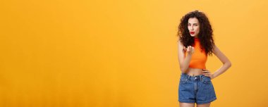 Kadın seksi erkeği baştan çıkarıyor. Şehvetli ve seksi, kıvırcık saçlı, modern kesimli bluzlu ve şortlu, çekici, çekici, çekici, turuncu arka planda flörtleşen kadın..