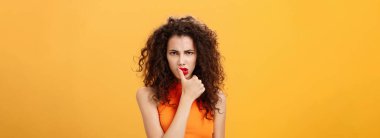 Ciddi kararlı ve odaklanmış, kıvırcık saçlı, kaşlarını çatan, tehlikeli, gözüpek, gözüpek, turuncu arka planda asi bir kadının belden yukarı çekimi. Vücut