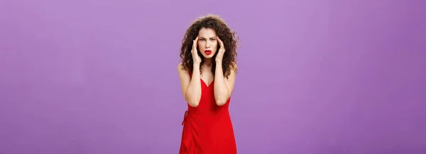 Stupid people getting on her nerves. Irritated and bothered stylish rish businesswoman in evening red dress pulling corners of eyes aside staring annoyed and unfocused at camera over purple background