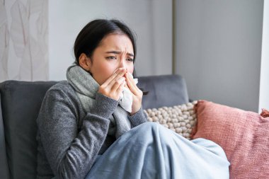 Hasta Koreli kadın resmi, sıcak kıyafetler ve atkıyla kaplı, hasta, üşütüyor ve kanepede hapşırıyor..