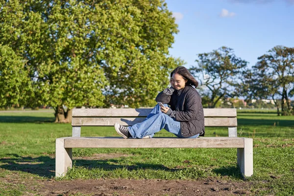 Young woman sitting alone on bench in park, using mobile phone, looking at screen, texting, sending messages in app chat.