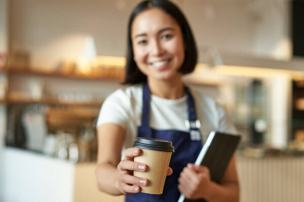 Blurred shot of young smiling barista girl, giving you takeaway cup of coffee, bring you takeout cappuccino and looking happy, working in cafe.