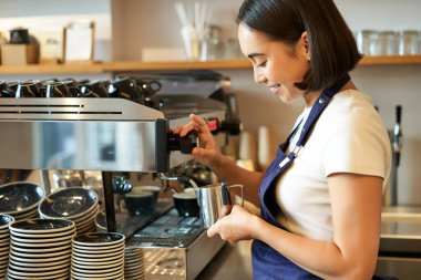 Gülümseyen Asyalı genç barista kız, kahve makinesi buharı kullanıyor, kapuçino siparişi için süt buharı yapıyor, önlüğün içinde duruyor, kafede çalışıyor..