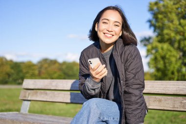 Elinde akıllı telefon olan, bankta oturan, gülümseyen, cep telefonu kullanan, parkta birini bekleyen hoş bir genç kadın..