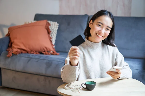 Young woman in living room, showing her credit card and using smartphone to pay, order online or shopping on application.