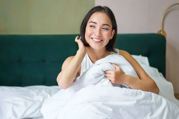 Close up of happy beautiful asian woman, waking up in bed and enjoys morning, looking outside window with sleepy smile on her face, hugging duvet.