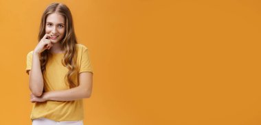 Yaratıcı, kurnaz, sarı tişörtlü, uzun saçlı, alnının altından şehvet ve niyetle bakan, parmağını ısıran, turuncu duvarın üzerinden kameraya gülümseyen genç bir kadın..