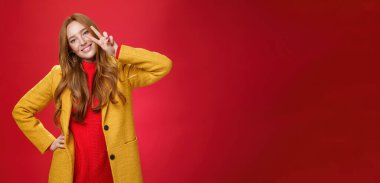 Mutlu bir şekilde gülümseyen, başını eğen ve yüzün yanında barış ya da zafer jesti gösteren zarif sarı ceket ve süveterle kırmızı arka planda poz veren arkadaş canlısı bir kız. Biçim