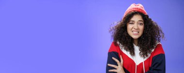 Kış şapkalı kıvırcık saçlı bir kadın rahatsız ve rahatsız hisseder dişlerini sıkarken ve kaşlarını çatarken kendine sarılırken kendine güvensiz ve garip davranır acımasız reddedilme demek istemez, garip hisseder.