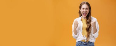 Güçlü hisseden kız neşelenmek ve kendine güveni arttırmak için bağırma sorununun üstesinden gelir neşeli ve zafer jestleriyle yumruklarını sıkarak bağırarak turuncu arka planda strese yol açar..