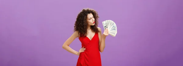 Woman deciding how spend lots of cash holding and looking at money. folding lips making decision or thinking how hard being rich standing elegant over purple background in red dress and curly