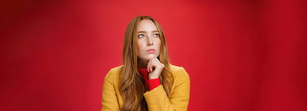 Close-up shot of determined and focused creative thoughtful redhead female looking at upper left corner touching chin, thinking, making choice or remembering information over red background.
